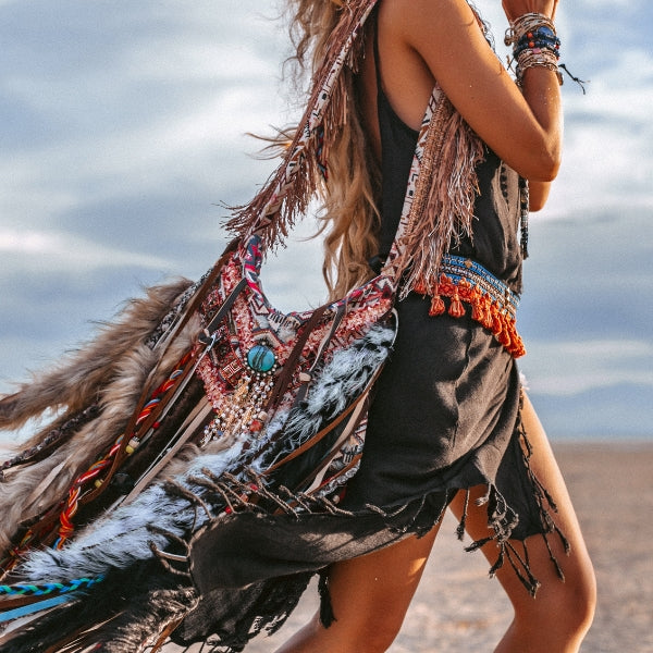 Boho girl with long hair, lots of bracelets carrying  a boho style bag with feathers, chains and beads