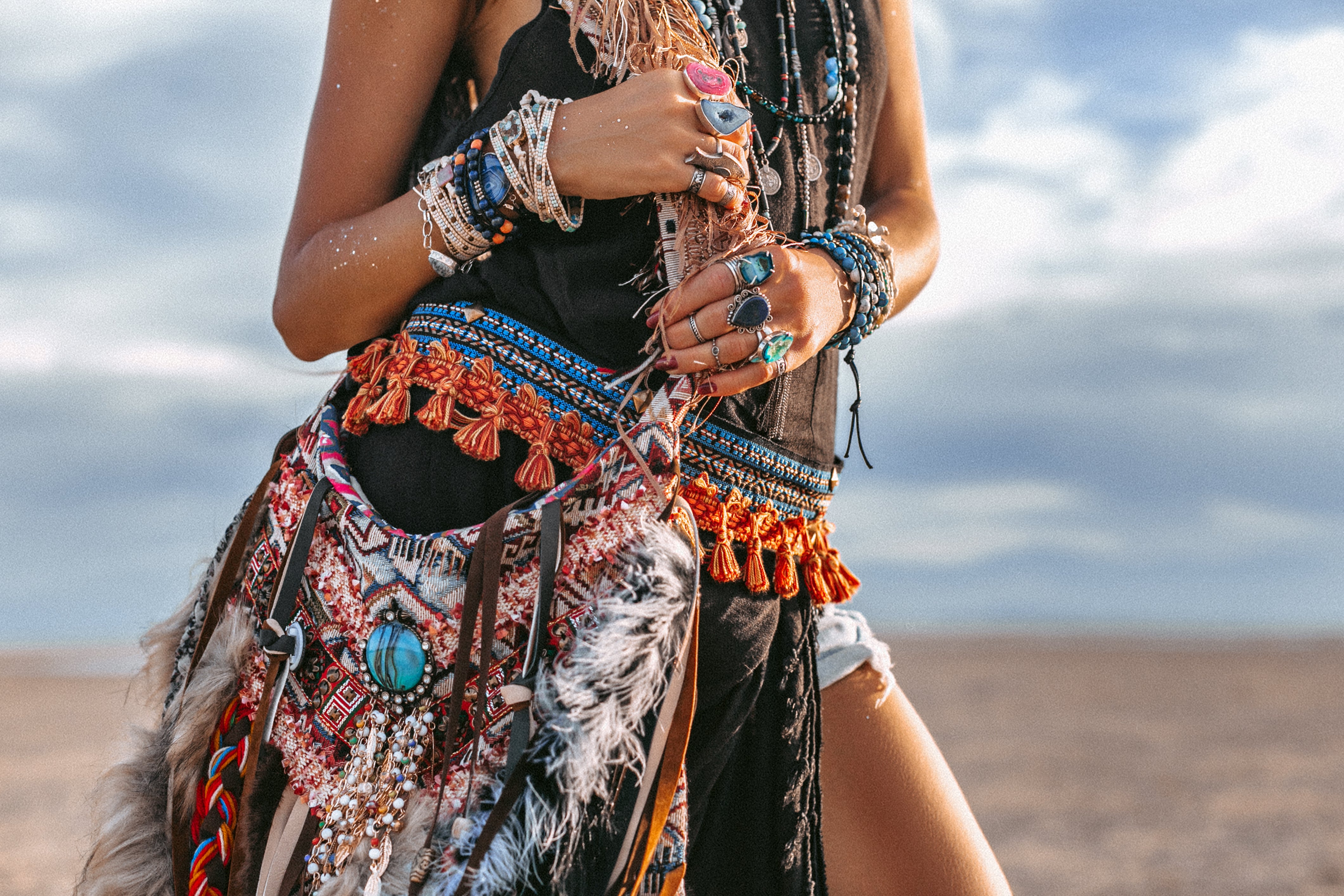 Boho girl with bracelets on her arms and rings on her fingers on beach carries a boho festival fringe bag 